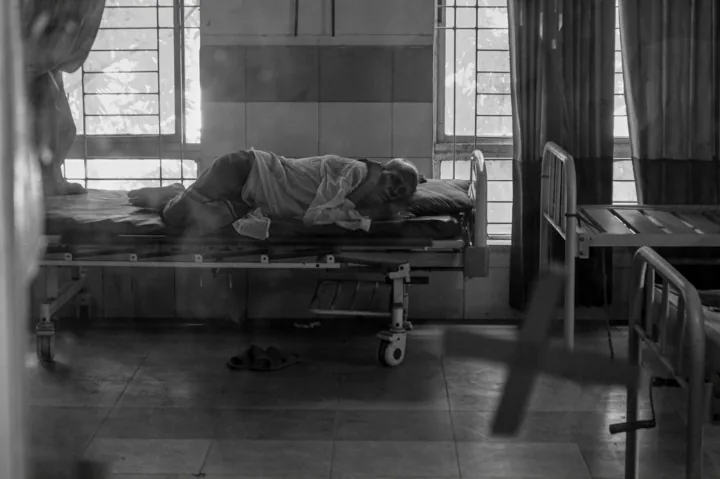 A patient rests curled on a hospital bed beside a sunlit window.