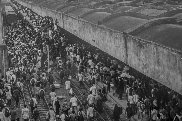 A dense crowd moves along railway tracks beside a long covered station platform.