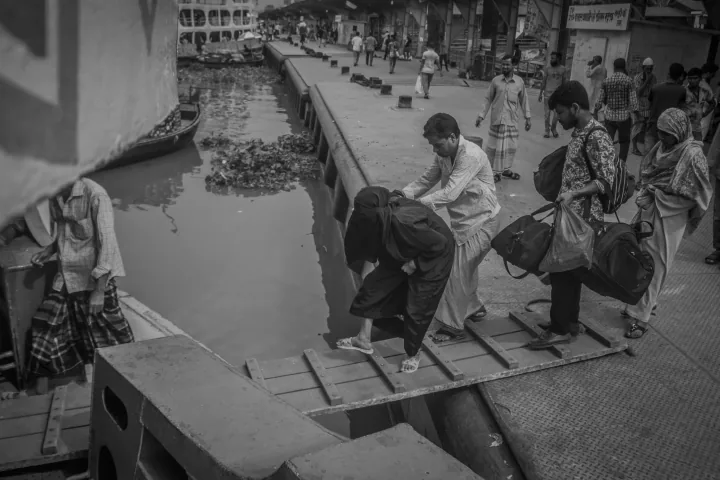A man helps a woman cross a narrow wooden gangplank while other passengers wait with bags.