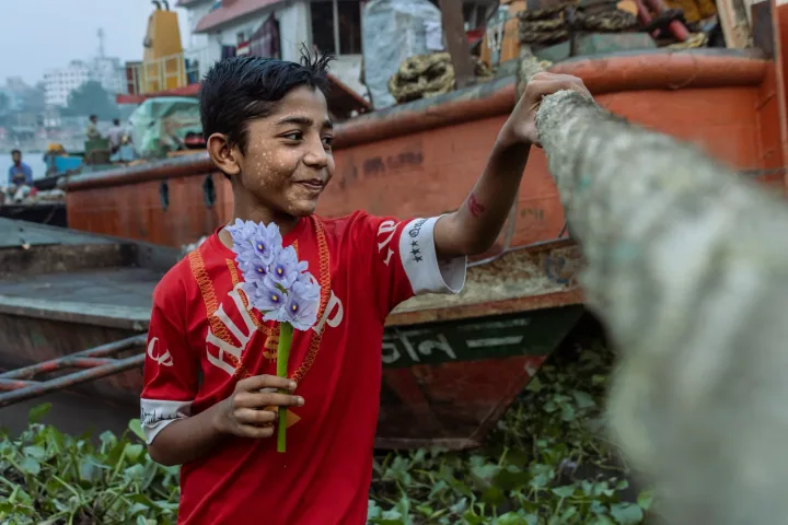 A smiling boy in a red shirt holds a water hyacinth flower beside moored boats.
