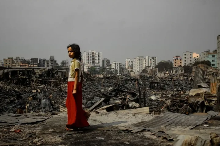 A young person stands among the burned remains of a settlement.