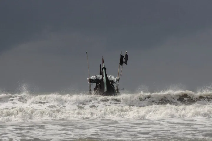 A fishing boat faces breaking waves beneath an overcast sky.