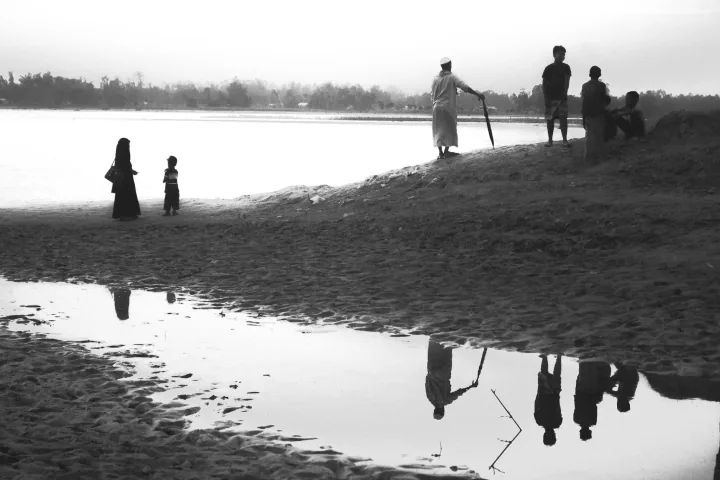 People stand along a riverbank with their silhouettes reflected in a pool in the foreground.