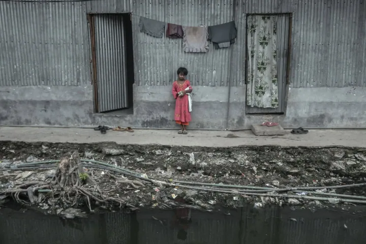 A child in a red dress stands outside a corrugated-metal home beside a littered waterway.