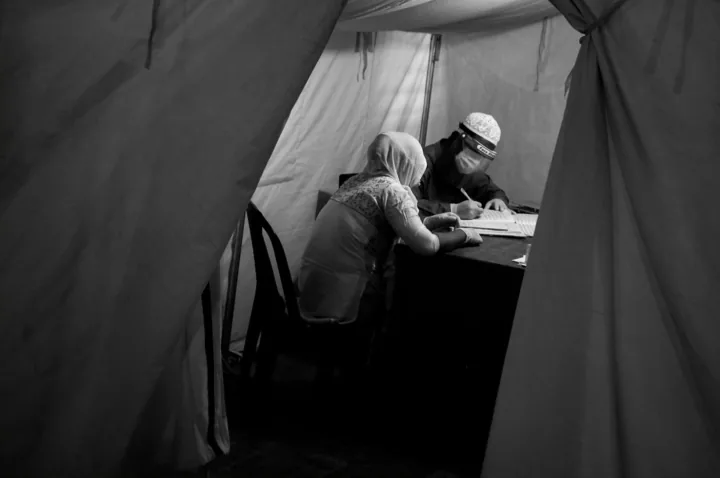 A masked clinician writes at a desk behind a makeshift cloth partition.