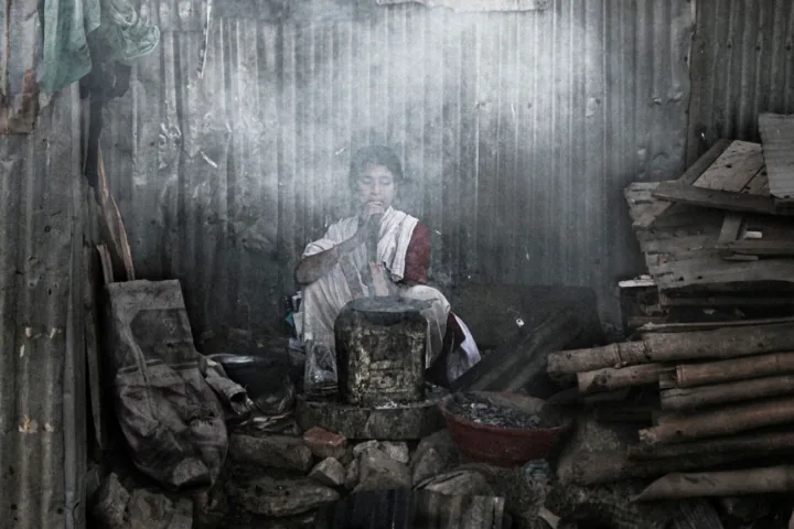 A woman tends a stove inside a smoky room with corrugated metal walls.
