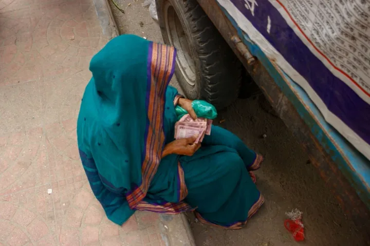 A woman in a turquoise sari counts banknotes beside the wheel of a bus.