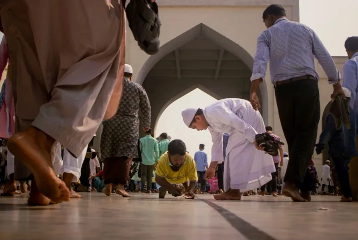A boy in white bends to give money to a man in a mosque courtyard as people pass.