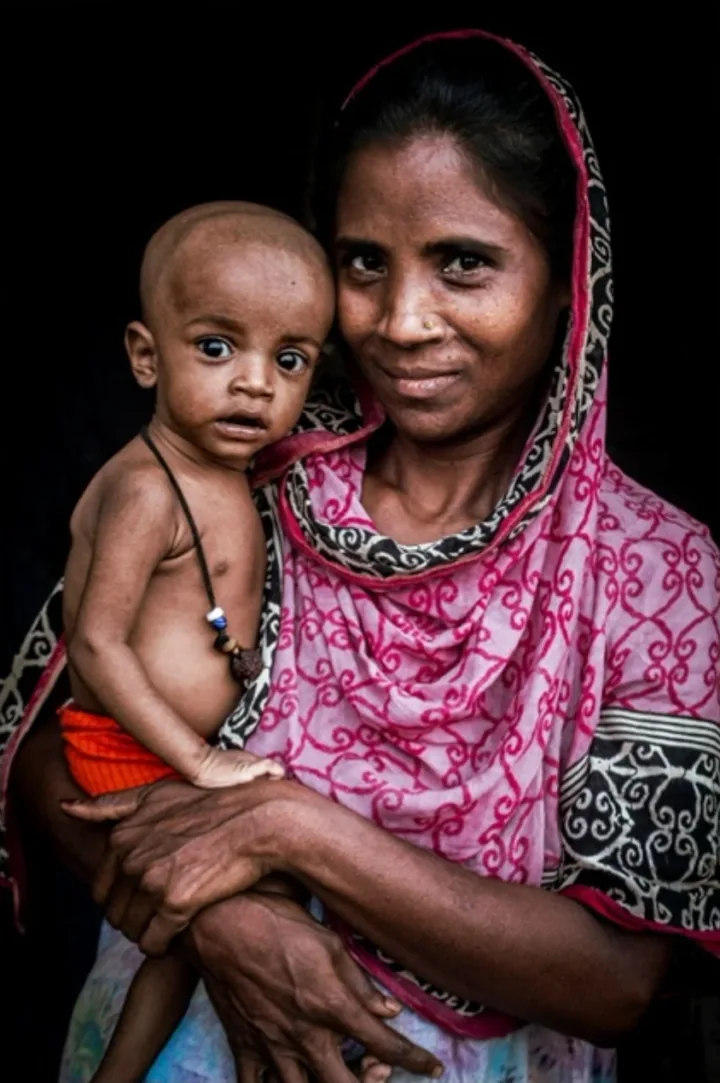 A woman in a pink patterned head covering holds her infant in a dark doorway.