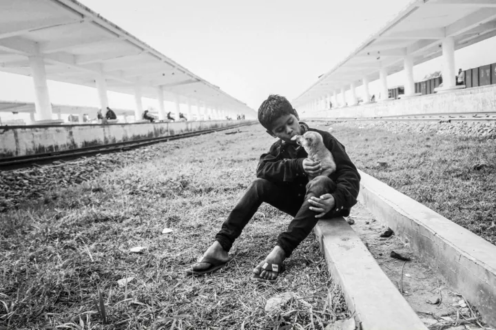 A boy sits beside railway tracks and holds a small puppy close to his face.