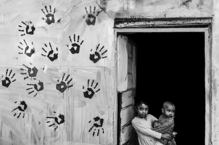 A girl holds a baby in a dark doorway beside a wall covered with handprints.