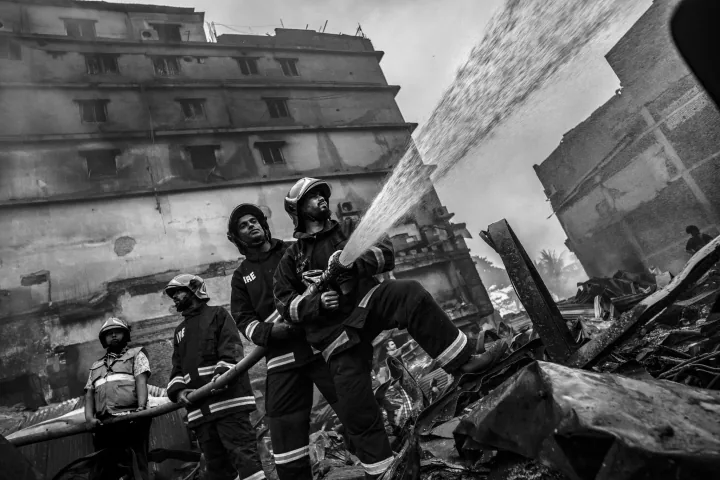 Helmeted firefighters work together to aim a hose amid damaged structures and debris.