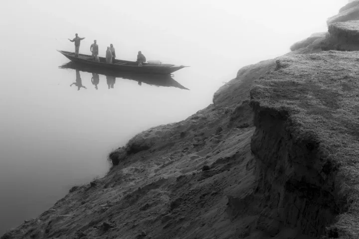 A narrow boat carrying several people floats on still, misty water beside a sloping riverbank.