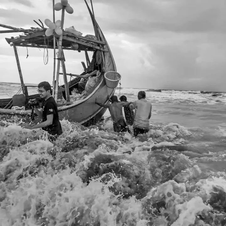 People stand beside a wooden fishing boat as waves break around their legs.