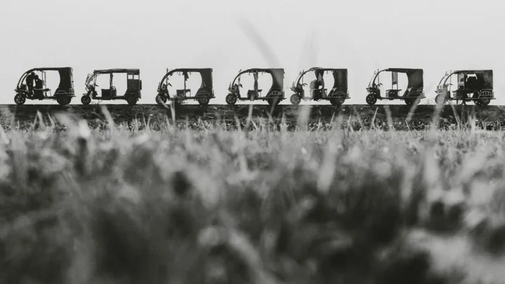 A row of auto rickshaws waits on a raised road beyond blurred foreground grass.