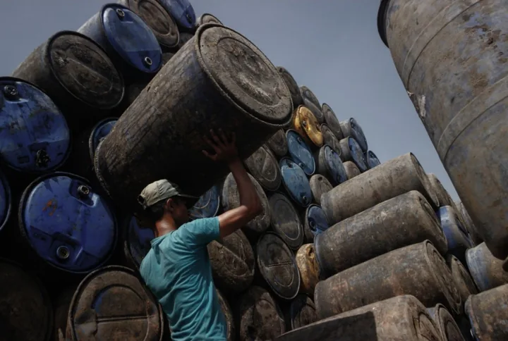 A worker lifts a large drum amid towering stacks of barrels.