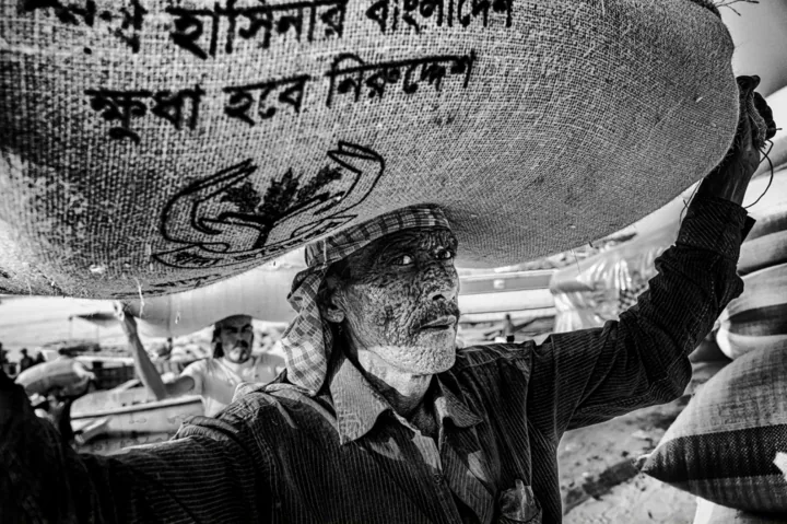 A porter carries a large grain sack printed with a Bangla slogan.