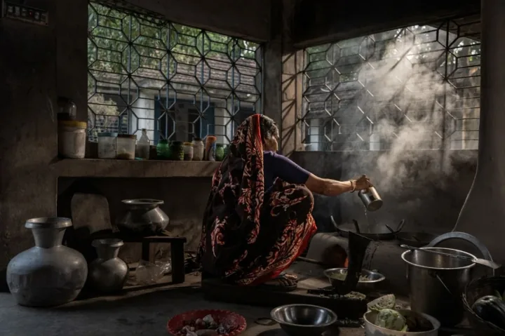A woman cooks beside a clay stove as sunlight streams through a kitchen window.