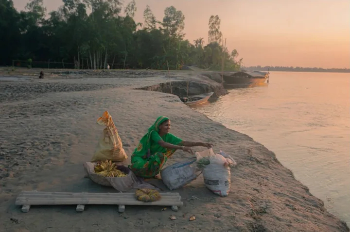 A woman sits beside baskets and sacks of produce on a riverbank at sunset.