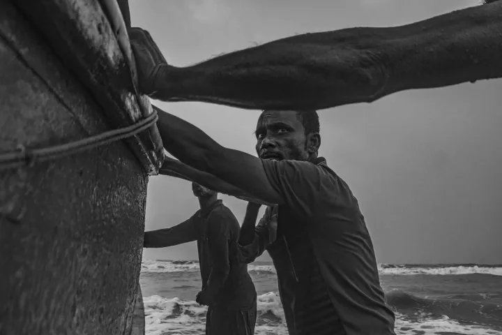 Men lean against the side of a fishing boat in the surf, photographed close to the hull.