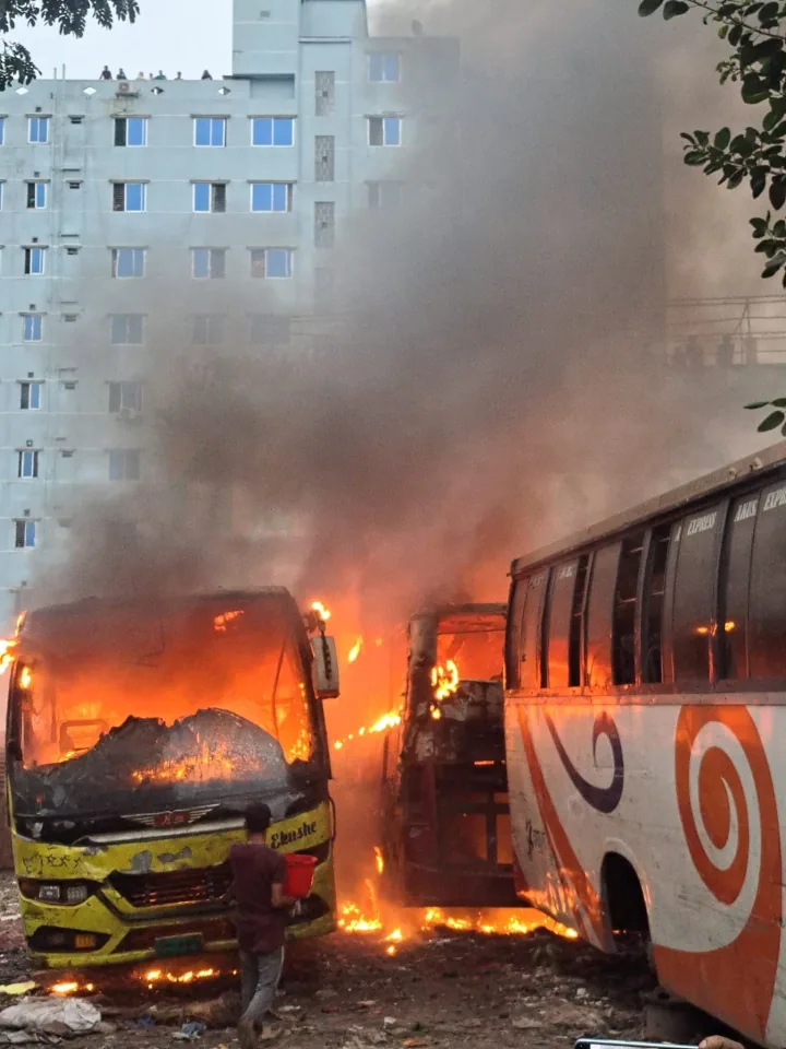 A bus burns beside another bus, sending thick smoke up in front of a tall building.