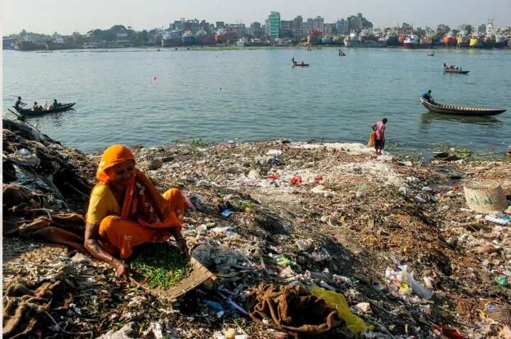 A woman in orange sorts green chillies on a rubbish-strewn riverbank.