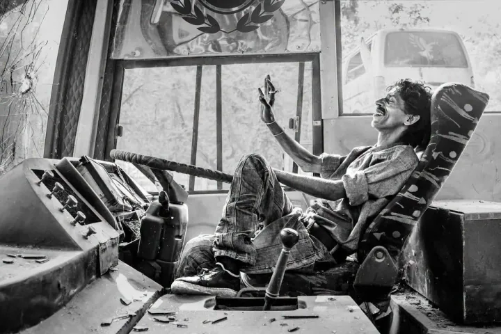 A man sits in a worn bus driver’s seat, smiling and raising one hand beside a damaged dashboard.