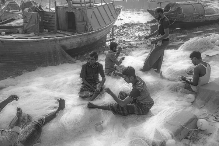 A group of people work with pale fishing nets spread across a riverfront landing beside wooden boats.