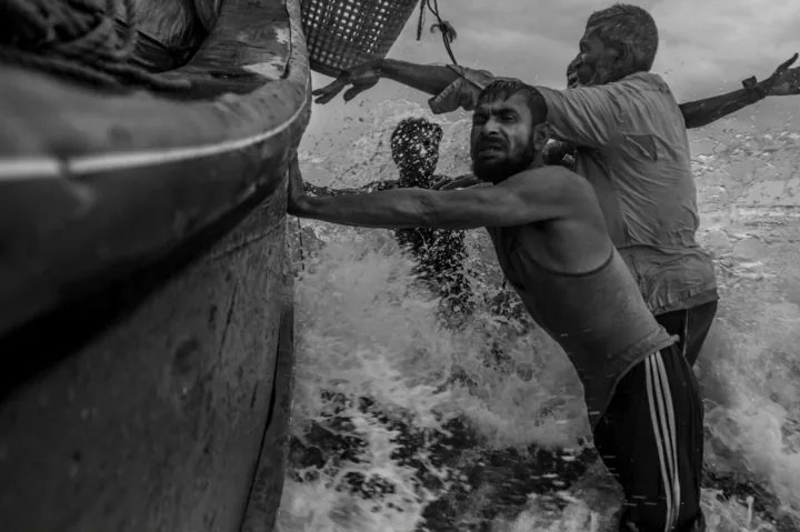 Men brace against a fishing boat as waves break around them.