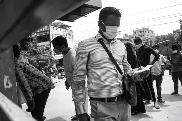 A masked man reads a paper among people waiting outside a hospital.