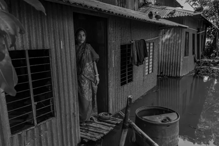 A woman stands at the doorway of a corrugated tin home surrounded by floodwater.