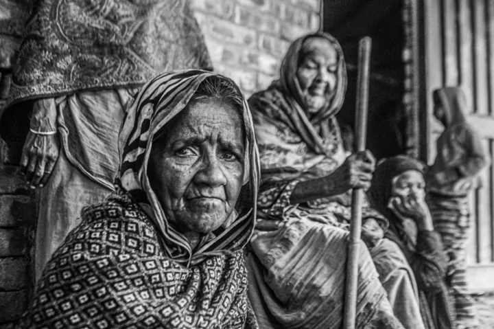 An elderly woman in a patterned head covering sits with other women against a brick wall.